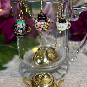 Può includere: Tre infusori da tè decorativi con disegni di gatti. Ogni infusore presenta un piccolo gatto all'interno di una tazza da tè, sospeso da una catena d'oro. Le tazze da tè sono decorate con motivi floreali e la parola "cafe". Un infusore a forma di teiera dorata è visibile sotto.