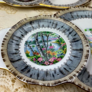 May include: A set of vintage porcelain serving dishes. The dishes feature a central landscape scene with trees, water, and flowers. The rims have a gray and brown pattern, with a white scalloped edge and gold trim. The dishes are on a cream-colored lace tablecloth.