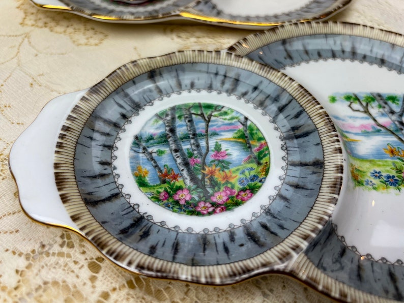 May include: A set of vintage porcelain serving dishes. The dishes feature a central landscape scene with trees, water, and flowers. The rims have a gray and brown pattern, with a white scalloped edge and gold trim. The dishes are on a cream-colored lace tablecloth.