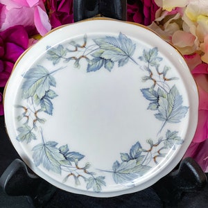 May include: White ceramic plate with a gold rim and a scalloped edge. The plate features a delicate design of blue and green leaves and seed pods arranged in a circular pattern. The plate is likely for serving or display.