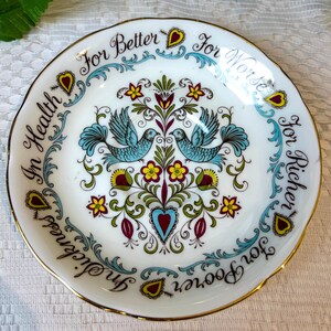May include: A white saucer with a gold rim features a blue and green floral design with two blue birds. The rim of the saucer is decorated with the text "For Better For Worse For Richer For Poorer In Health In Sickness."