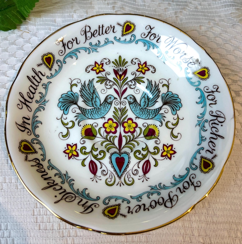 May include: A white saucer with a gold rim features a blue and green floral design with two blue birds. The rim of the saucer is decorated with the text "For Better For Worse For Richer For Poorer In Health In Sickness."