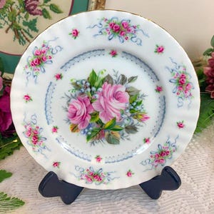 Puede incluir: Plato de porcelana blanca con borde dorado, adornado con rosas rosas, lazos azules y hojas verdes. El plato tiene un borde festoneado y está sobre un soporte negro. Diseño floral decorativo.