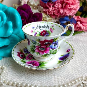 May include: A fine bone china teacup and saucer set, decorated with a floral pattern in shades of red, pink, and purple. The word "March" is written on the cup. The cup has a gold handle and trim.