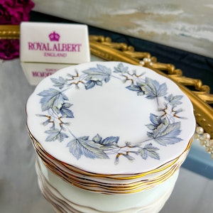 May include: A stack of six white ceramic dessert plates with a blue and gold floral pattern. The plates feature a gold rim and the words "Royal Albert England" on the top plate.