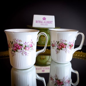 Puede incluir: Dos tazas de té blancas con bordes dorados y patrones florales. Las tazas están en una bandeja dorada con una caja verde que dice "Royal Albert England".