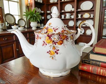 Pode incluir: Bule de porcelana branco com um design floral com rosas amarelas e vermelhas. O bule tem uma alça e acabamentos dourados e uma tampa a condizer. O bule está sobre uma mesa de madeira, com livros e outros utensílios de cozinha no fundo.