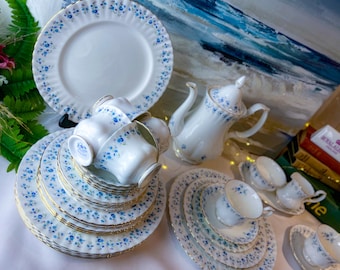 Puede incluir: Un juego de tazas de té y platillos blancos con un patrón floral azul. El juego incluye una tetera y un plato más grande. Las tazas de té y los platillos están apilados uno encima del otro.