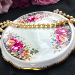 May include: A white porcelain trinket dish with a gold rim and a floral design. The dish has a scalloped edge and is decorated with pink, yellow, and orange roses. A strand of pearls is draped around the dish.