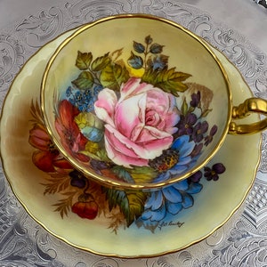 Op de afbeelding: Een vintage theekop en schotel set. De lichtgele theekop en schotel hebben een gouden rand en een bloemenontwerp met een grote roze roos en andere kleurrijke bloemen. De schotel is gesigneerd "L.A. Bailey".