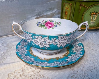 May include: A turquoise and white floral patterned teacup and saucer set. The cup has two handles and a floral design with a pink rose in the centre. The saucer has a matching floral design.