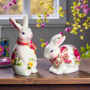 May include: Two white ceramic bunny figurines with floral designs. One bunny stands upright, the other is seated. Both have pink accents, blue eyes, and decorative collars. The bunnies are decorated with red, yellow, and pink flowers.