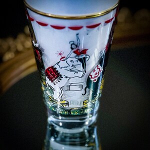 May include: A clear glass shot glass with a gold rim and a circus-themed design. The design features a white elephant, a red and white striped tent, and a black and white figure performing on a trapeze.