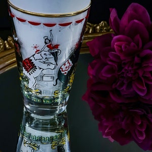 May include: A vintage glass with a white background and a gold rim. The glass features a colorful illustration of a circus scene with an elephant, a performer, and a tent. The glass is reflected in a mirror with a gold frame.