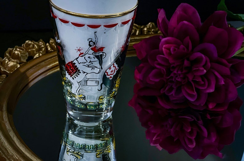 May include: A vintage glass with a white background and a gold rim. The glass features a colorful illustration of a circus scene with an elephant, a performer, and a tent. The glass is reflected in a mirror with a gold frame.