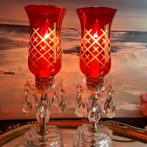 Może przedstawiać: Para czerwonych szklanych świeczników z wiszącymi kryształowymi pryzmami. Świeczniki mają biały grawerowany wzór na szkle.