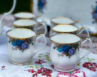 May include: A set of white teacups with blue and purple floral designs and gold trim. The teacups are arranged on a white tablecloth with a floral pattern.