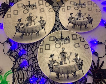 May include: Three white ceramic dinner plates with black and white illustrations of skeletons having tea. The plates have a black and white pattern of squares and a chandelier. The plates are on a black spiderweb tablecloth with purple fairy lights.