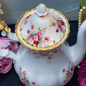 Puede incluir: Una tetera de porcelana blanca con ribete dorado y un patrón floral. La tetera tiene una tapa con un diseño floral y un ribete dorado. La tetera está sobre una superficie negra.