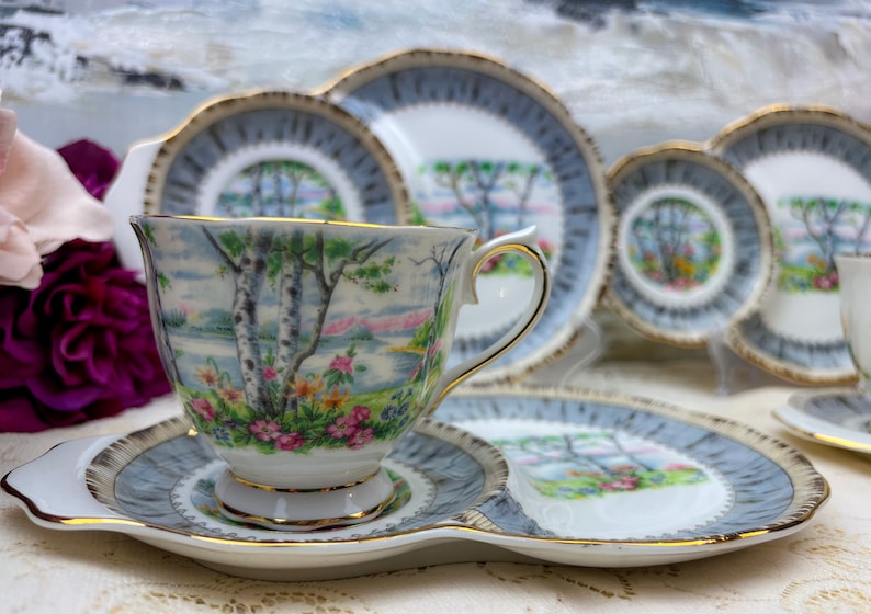 May include: A vintage tea set featuring a cup, saucer, and plates with a landscape design. The cup and plates have a light blue border with gold trim. The cup and plates feature a landscape scene with trees and flowers.