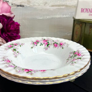 May include: A set of three white porcelain soup bowls with a delicate pink floral pattern and gold trim. The bowls are stacked on top of each other. The pattern features pink roses and green leaves.