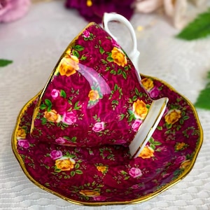 Puede incluir: Un juego de taza y platillo de té vintage con un estampado floral. El juego presenta un fondo burdeos oscuro adornado con rosas amarillas y rosas, acentuado con ribetes dorados. La taza descansa sobre su platillo, con un asa blanca y una cuchara a juego.