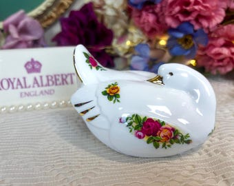 May include: A white porcelain swan figurine with gold accents and floral rose details. The Royal Albert England logo is visible in the background. The swan has a glossy finish and is decorated with pink, yellow, and green roses.