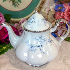 Peut inclure: Théière en porcelaine blanche avec garniture et anse argentées. La théière présente un délicat motif floral bleu sur sa surface et sur le couvercle. Le bec et la poignée sont rehaussés d'argent.