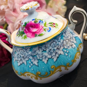 Op de afbeelding: Een vintage porseleinen suikerpot met deksel. De kom is turquoise met witte bloempatronen en gouden accenten. Het deksel heeft een roze roos en andere bloemendetails. De handgreep is wit met gouden rand.