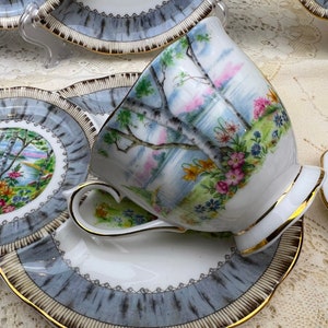May include: A vintage teacup and saucer set. The white porcelain cup and saucer feature a landscape scene with trees and flowers. The rims are trimmed with gold and a gray and white pattern. The set is designed for tea service.