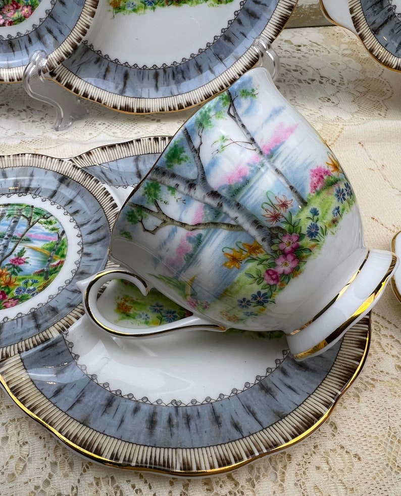 May include: A vintage teacup and saucer set. The white porcelain cup and saucer feature a landscape scene with trees and flowers. The rims are trimmed with gold and a gray and white pattern. The set is designed for tea service.