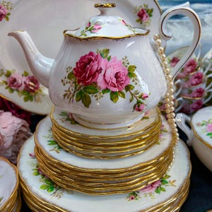 Może przedstawiać: Biały porcelanowy zestaw do herbaty z różowymi wzorami kwiatowymi róż i złotymi obramowaniami. Zestaw zawiera imbryk, ułożone w stos spodki i półmisek. Perłowy naszyjnik jest przewieszony przez uchwyt imbryka.