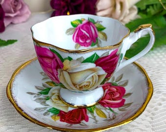 May include: A white teacup and saucer set with a floral rose design. The teacup and saucer are trimmed with gold and feature pink, red, and yellow roses with green leaves. The teacup has a gold handle.