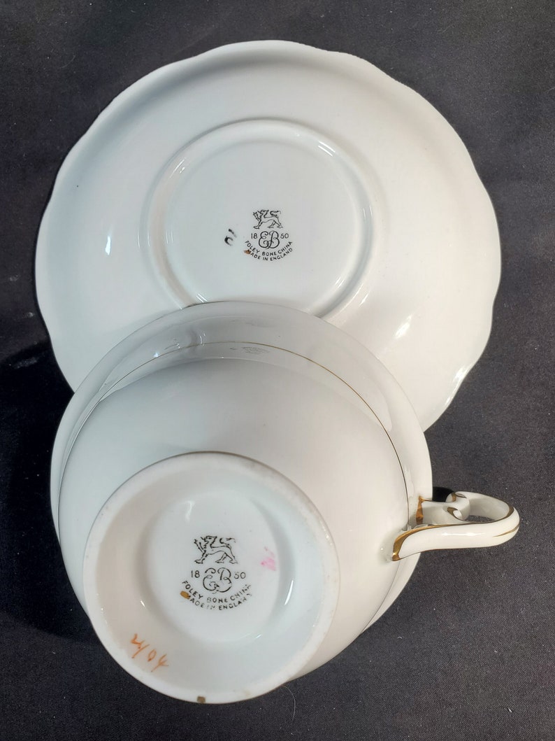 Puede incluir: Taza de t&eacute; y platillo blancos con ribete dorado. La parte inferior de la taza y el platillo tienen un logotipo en blanco y negro que dice "18 R 50 POLEY BONE CHINA MADE IN ENGLAND".