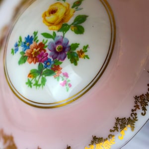 Puede incluir: Una taza de t&eacute; de porcelana rosa con un dise&ntilde;o floral blanco y ribete dorado. El dise&ntilde;o floral presenta una rosa amarilla y otras flores de colores.