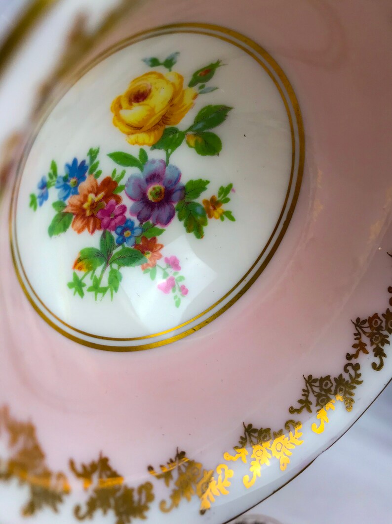 Puede incluir: Una taza de t&eacute; de porcelana rosa con un dise&ntilde;o floral blanco y ribete dorado. El dise&ntilde;o floral presenta una rosa amarilla y otras flores de colores.