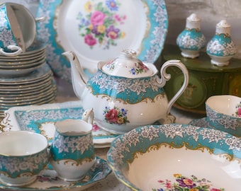 Pode incluir: Um conjunto de chá de porcelana vintage. O conjunto inclui uma chaleira, chávenas, pires, pratos e travessas. As peças são brancas com um design floral turquesa e detalhes dourados. A chaleira tem aproximadamente 18 cm de altura.