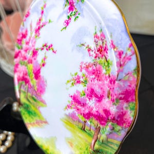 May include: An oval decorative dish with a hand-painted landscape of pink blossom trees, green grass, and a pale blue sky. The dish has a gold trim and a scalloped edge, giving it a vintage appearance.