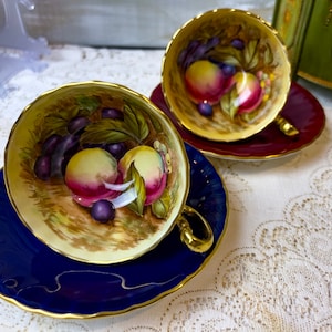 May include: Two teacups and saucers with a floral design. The teacups have a cream-colored background with a painted design of peaches, plums, and leaves. The saucers are blue and gold.