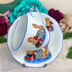 May include: A white teacup and saucer set with a blue trim. The set features a whimsical illustration of children playing in the water with dolphins and toy horses. The cup is perched on the saucer, which has a scalloped edge.