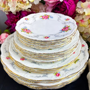 Puede incluir: Una pila de platos de porcelana blanca con borde dorado y un diseño floral con rosas rosas y azules. Los platos tienen un borde festoneado y un borde decorativo gris.