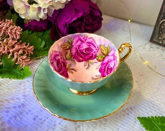 Può includere: Un set vintage di tazza da tè e piattino con un design floreale. La tazza è bianca con rose rosa e foglie verdi. Il piattino è di colore verde turchese chiaro con un bordo dorato.