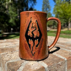 May include: A brown ceramic mug with a black and gold dragon symbol. The mug has a rustic, hand-crafted look.