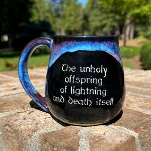 May include: A black ceramic mug with a blue and purple glaze. The mug has a white inscription that reads "The unholy offspring of lightning and death itself."