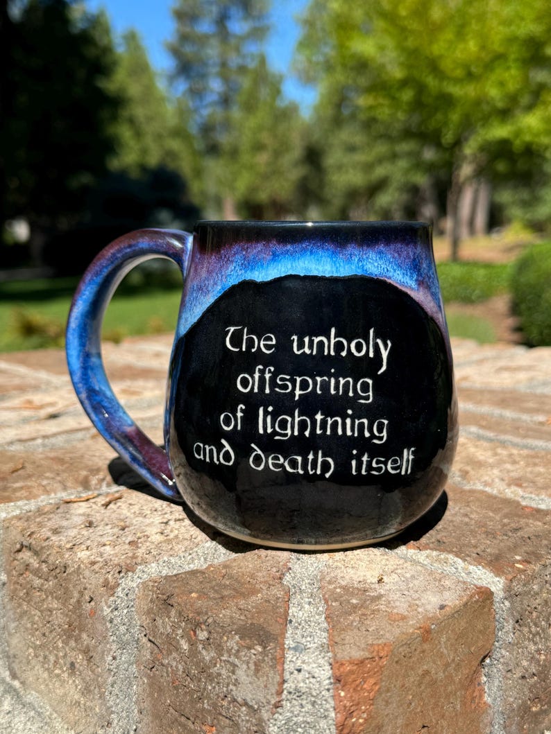 May include: A black ceramic mug with a blue and purple glaze. The mug has a white inscription that reads "The unholy offspring of lightning and death itself."