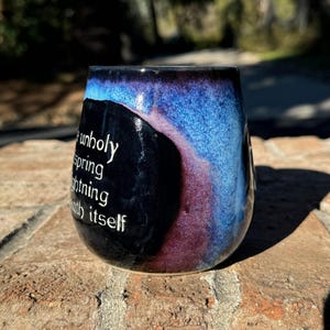 May include: A black ceramic mug with a blue and purple glaze. The mug has a quote on the side that reads "an unholy spring lightning with itself".