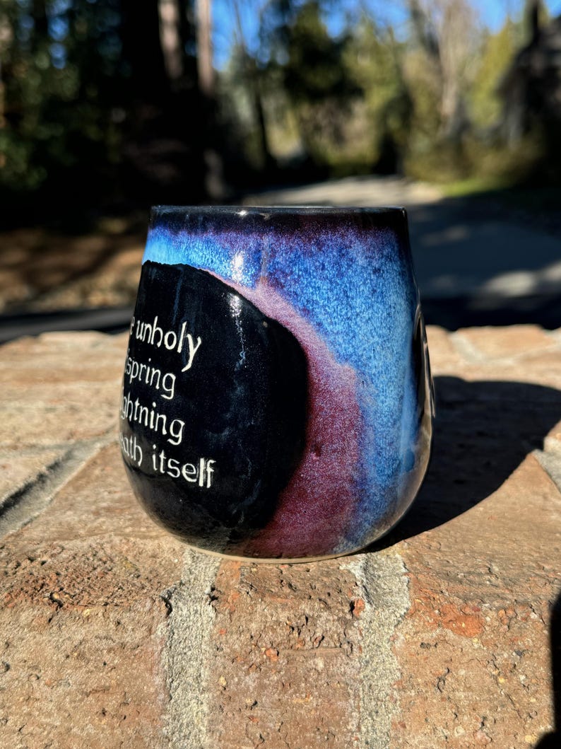 May include: A black ceramic mug with a blue and purple glaze. The mug has a quote on the side that reads "an unholy spring lightning with itself".
