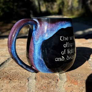 May include: A black ceramic mug with a blue and purple glaze. The mug has a handle and a quote on the side that reads "The un- offspring of light and death."