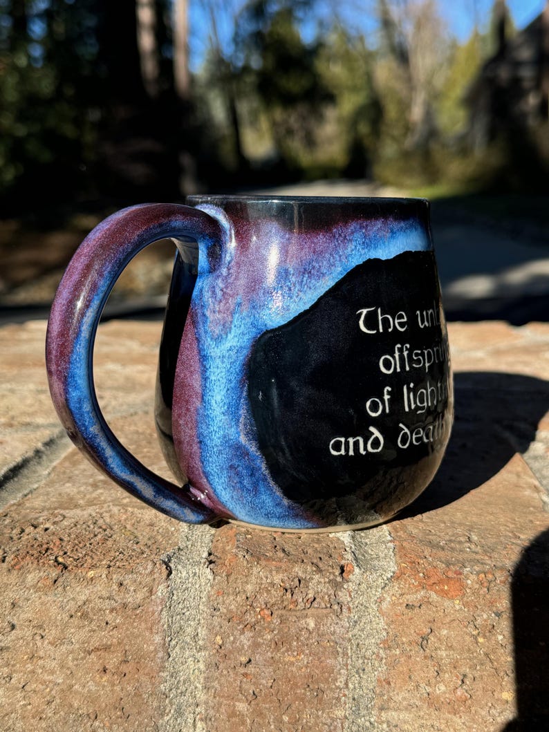 May include: A black ceramic mug with a blue and purple glaze. The mug has a handle and a quote on the side that reads "The un- offspring of light and death."