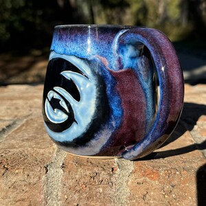 May include: A blue and purple ceramic mug with a black and white swirl design. The mug has a handle and is sitting on a brick surface.